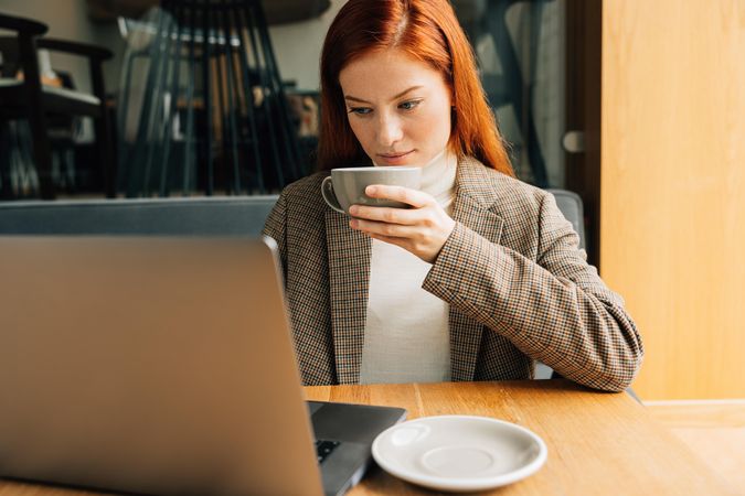 Red haired woman sipping coffee while sitting at laptop