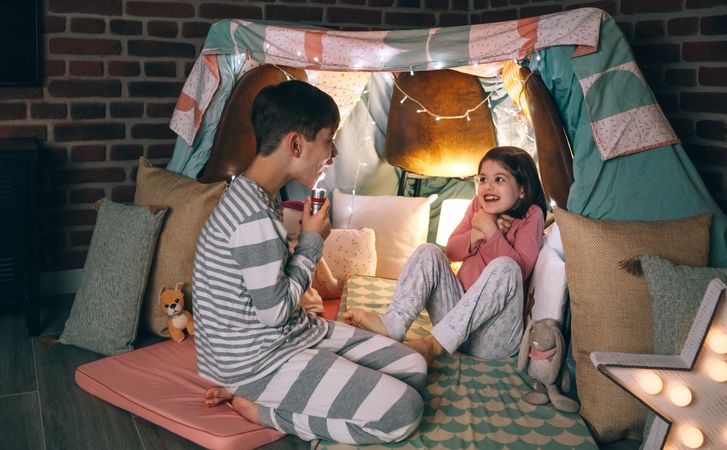 Happy little girl shrinking and shouting while brother making scary face illuminated with lantern