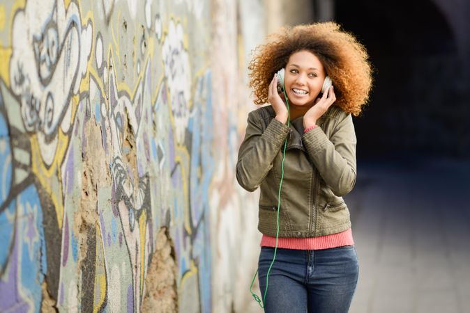 Smiling woman with curly hair and green jacket pictured using headphones in backstreet