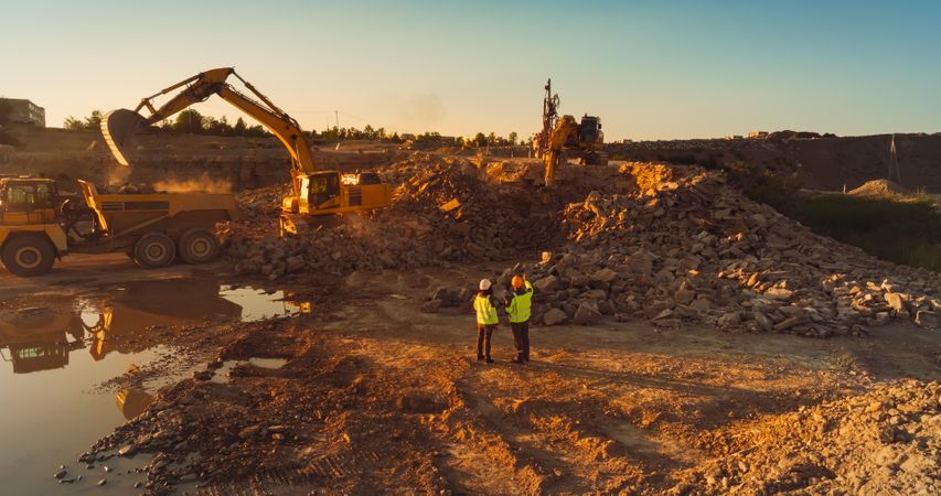 Aerial Drone Shot Of Construction Site On Sunny Day: Industrial Excavators Digging Rocks And Loading Into A Truck. Engineer And Inspector Observing Process, Discussing New Real Estate Project