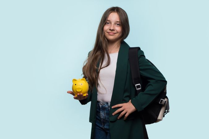 Teen girl holding yellow piggy bank, wearing backpack, smiling confidently.