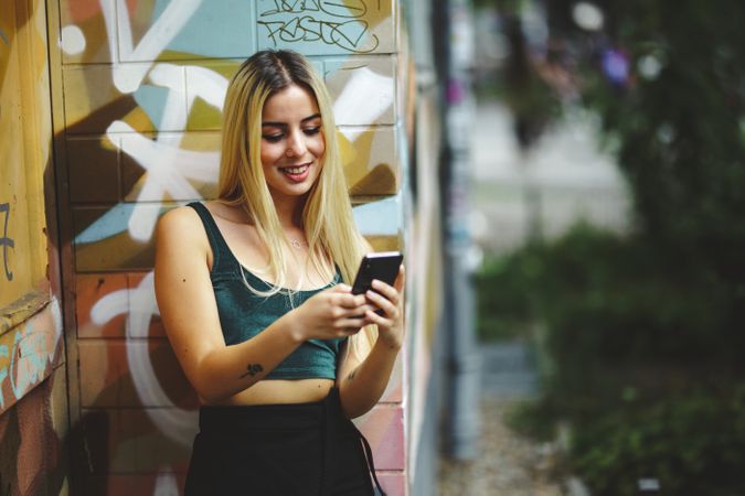 Stylish woman using smartphone at graffiti wall on city street.