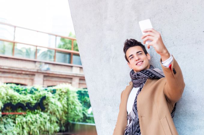 Male in camel coat and scarf standing outside taking selfie