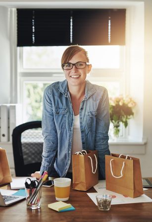 Smiling business woman at her home office with bags of product for clients, vertical