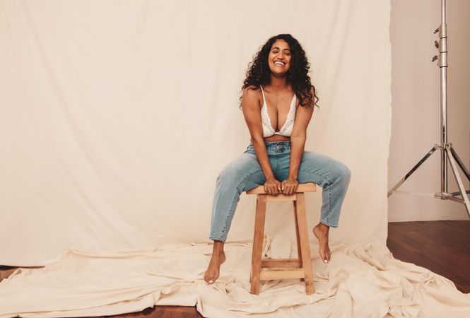Happy young woman sitting on a wooden chair while wearing jeans and a bra