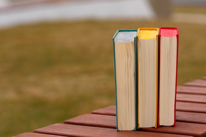 Three hardcover books standing vertically on wooden outdoor bench.