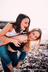 Two young woman having fun with one giving the other a piggyback on the beach v5lVYb