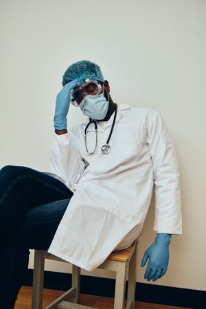 Tired Black male doctor in ppe gear sitting down in hospital with eyes closed