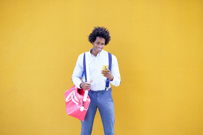 Man with curly hair wearing shirt and suspenders in front of yellow wall checking phone