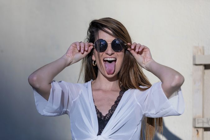 Young woman wearing sunglasses standing in the street while sticking tongue out to the camera showing on a sunny day