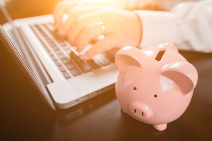 Piggy Bank Near Male Hands Typing on Laptop Computer.
