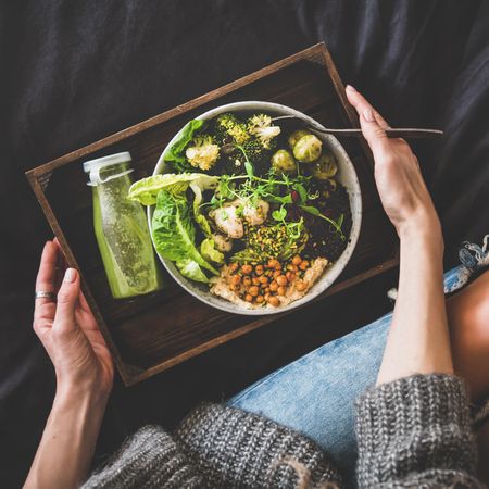 Woman in jeans sitting with wooden tray of vegetarian bowl with smoothie on side, square crop