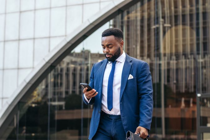 Black business man in suit leaving the office holding his work briefcase and using smartphone