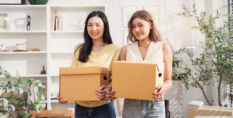 Happy Asian woman friends holding cardboard boxes moving to the new home