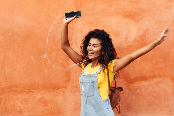 Dancing Arab woman with curly hairstyle using phone and headphones with hands in above her head