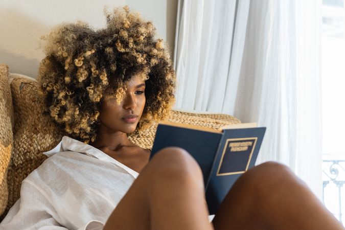 Young Black woman sitting on bed and reading book