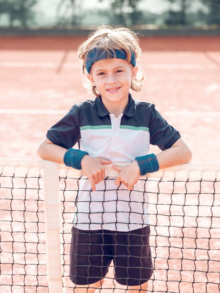 Cute sporty kid smiling at camera while standing on tennis court