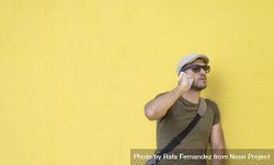 Male in hat and sunglasses standing next to yellow wall and speaking on phone, copy space 4dlMn4