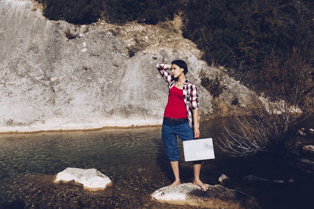 woman standing on rock with case near clear water in lake