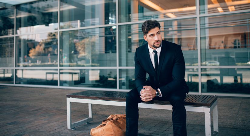Businessman sitting on bench with handbag and looking away