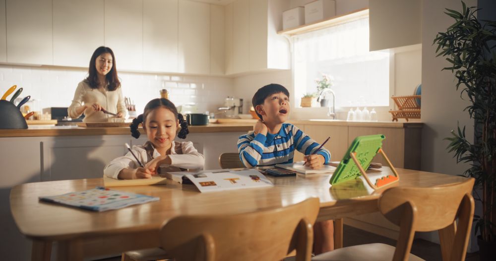 Portrait of a Korean Children Sitting at a Kitchen Table, Drawing and Writing While their Mother is Preparing Breakfast.