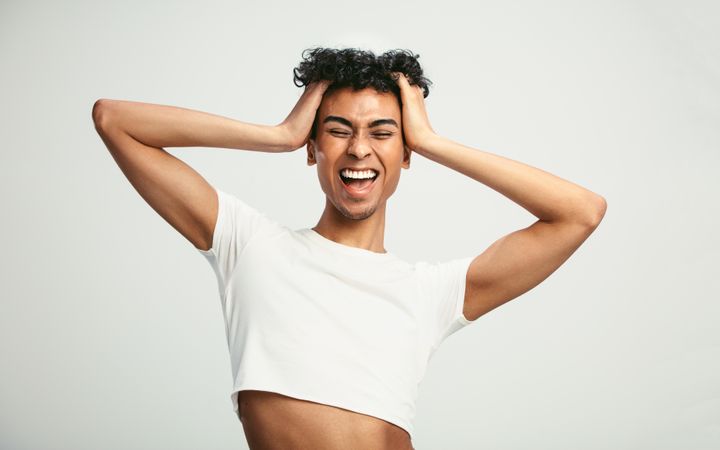 Man wearing crop top laughing with his hands in hair against light background
