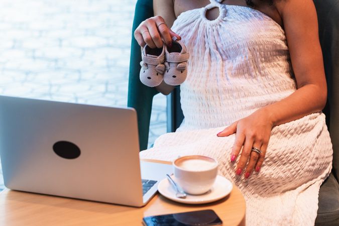 Anonymous pregnant Black woman showing baby booties to laptop