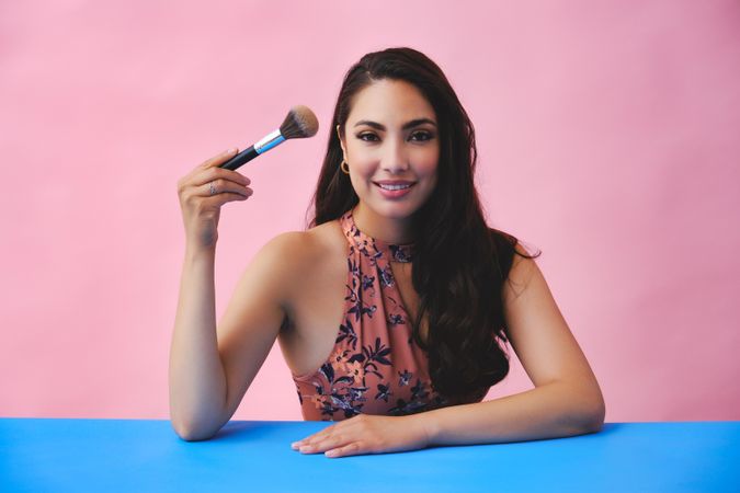 Happy Hispanic woman with long brown hair holding up large make up brush and looking at camera