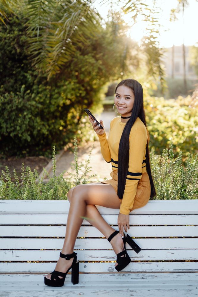 Charming long haired Asian woman with smartphone sitting on benc