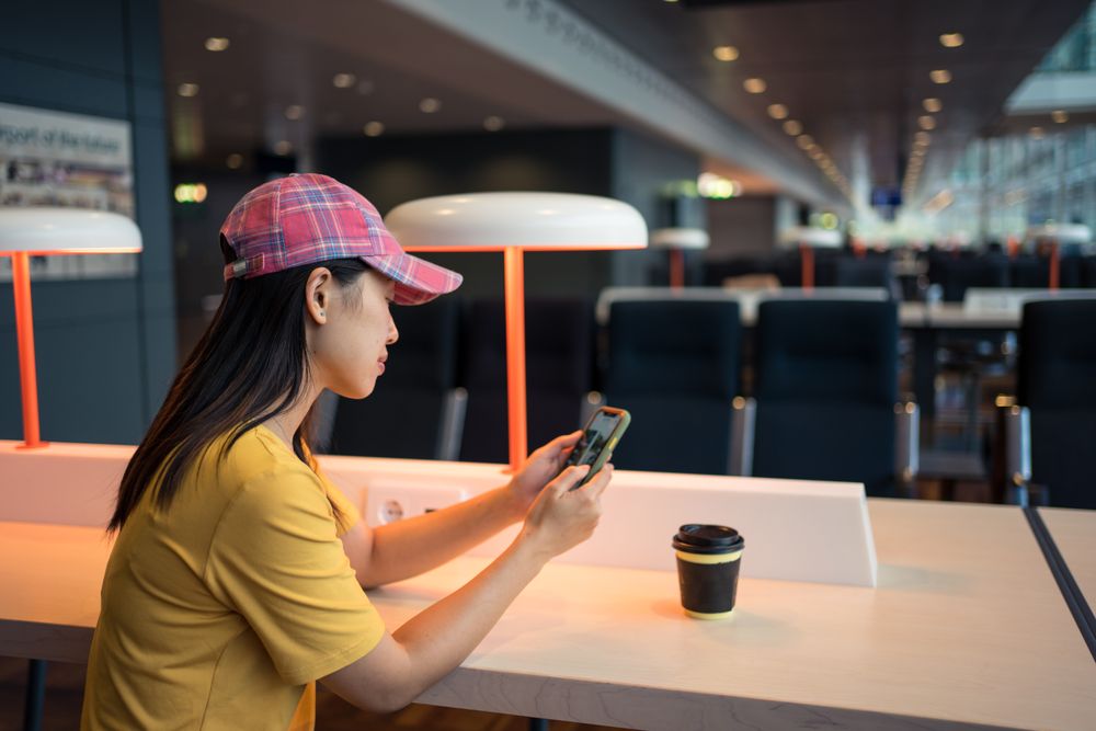 Asian woman browsing smartphone while drinking coffee at table under lighting lamp