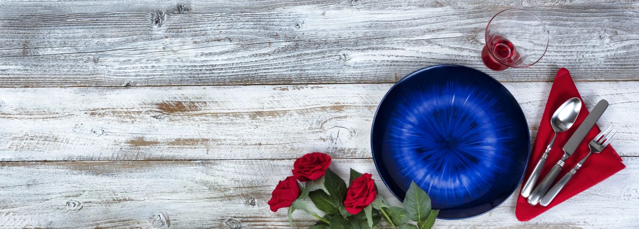 Table setting for Valentines Day celebration in flat lay format with blue plate, red roses, napkin, silverware and drinking glass on rustic wood