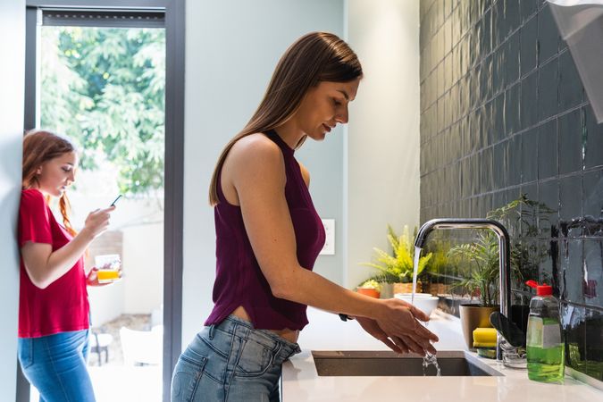 Woman washing hands in kitchen near friend using smartphone