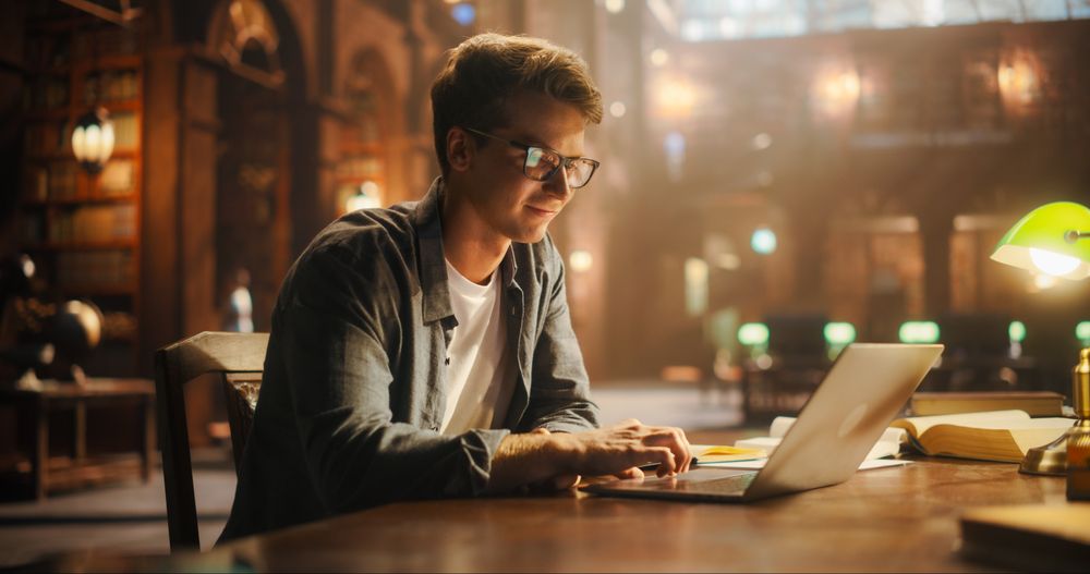 Focused Young Man Studying in a Classical Library, Using Laptop for Online Education. Male Engrossed in E-Learning