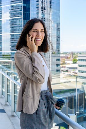 A woman on her office balcony talks on her cell phone and holds a paper cup of coffee during her break.