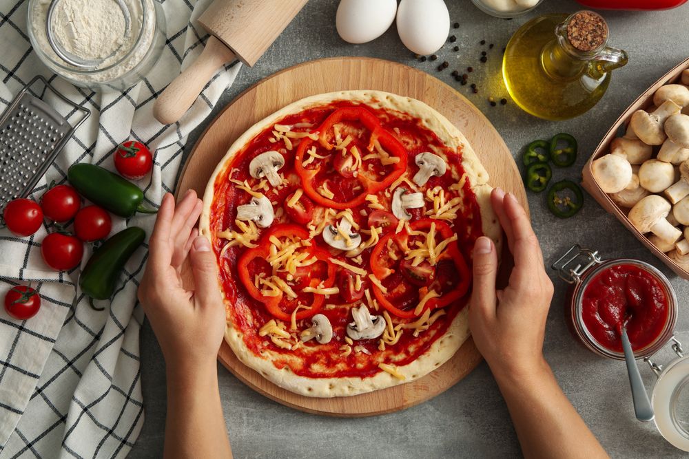 Female hands hold raw pizza on gray background with ingredients for cooking