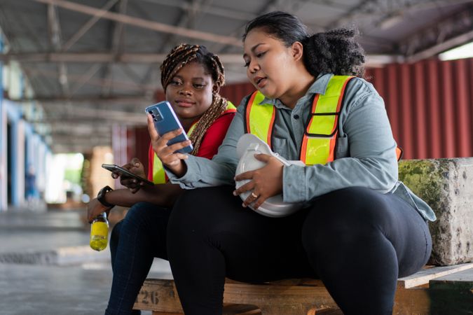 Two female workers take a break, sitting and watching on mobile phone.