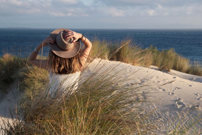 Woman in hat sitting on sandy hill enjoying sea view