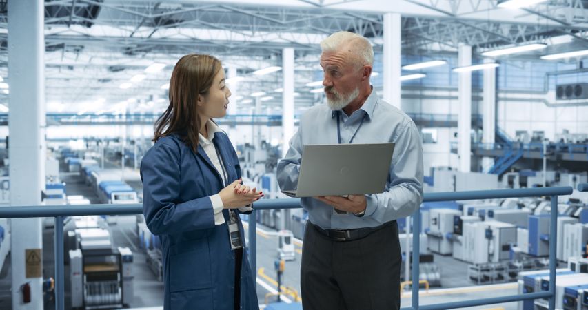Portrait of an Engineer and Happy Asian Scientist Using Laptop Computer and Talking in a Factory Facility with Equipment Producing Modern Electronic Components for Different Industries
