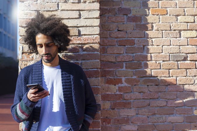 Man looking at his smartphone while leaning on a brick wall outdoors on sunny day with copy space