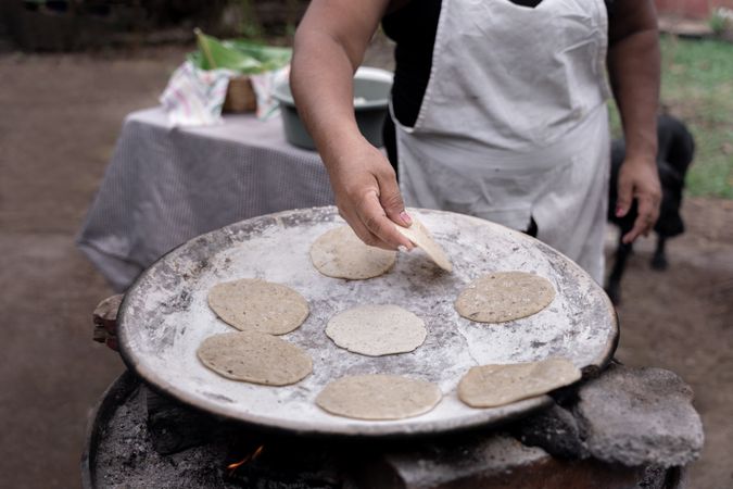 An adult Hispanic woman is cooking traditional corn tortillas over an outdoor stove