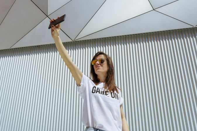 Cheerful woman taking selfie and posing near modern wall