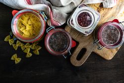 Top view of ingredients in glass containers with farfelle pasta, seeds and beans on wooden background with copy space