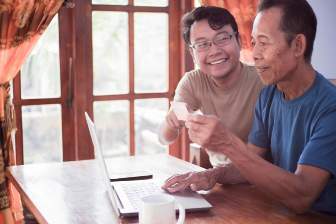 Happy adult son and older father pay online using laptop and credit card