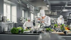 Famous Chef Works in a Big Restaurant Kitchen with His Help. Kitchen is Full of Food, Vegetables and Boiling Dishes. He is trying taste.
