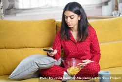 Female in red pajamas sitting on sofa with bowl of chips and remote control 56qGY0