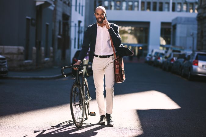 Portrait of man walking with bicycle to work and talking on phone
