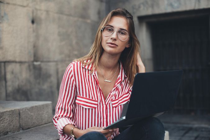 Fashionable young woman with glasses sitting on outdoor steps in European city with laptop