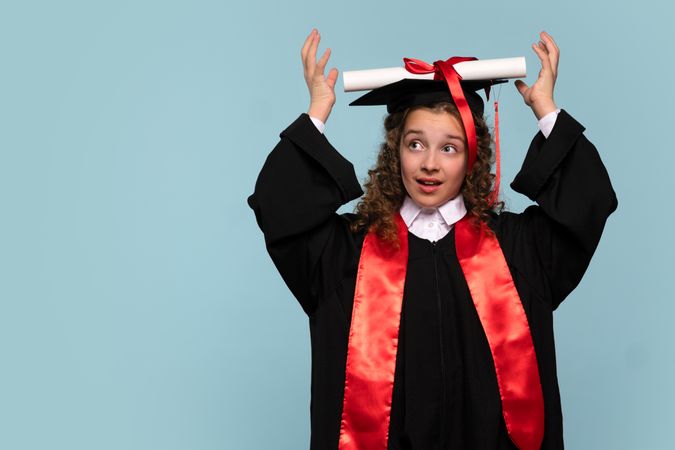 Young girl in graduation gown placing diploma on head playfully.