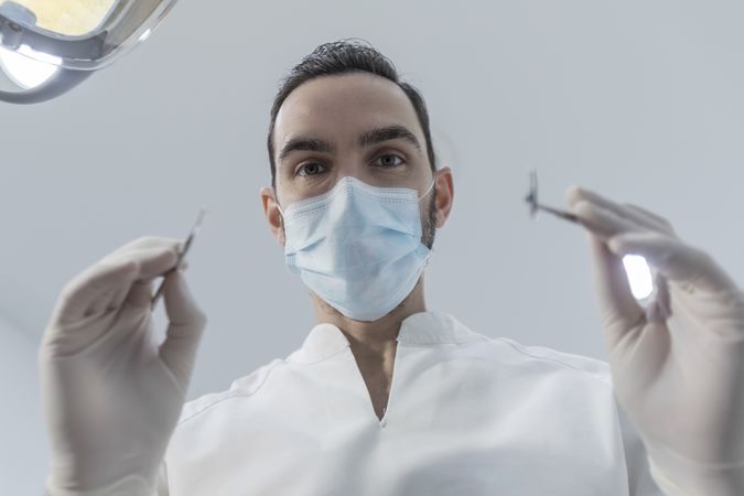 Low angle view of male dentist in medical mask holding dentist tools for dental procedure in clinic