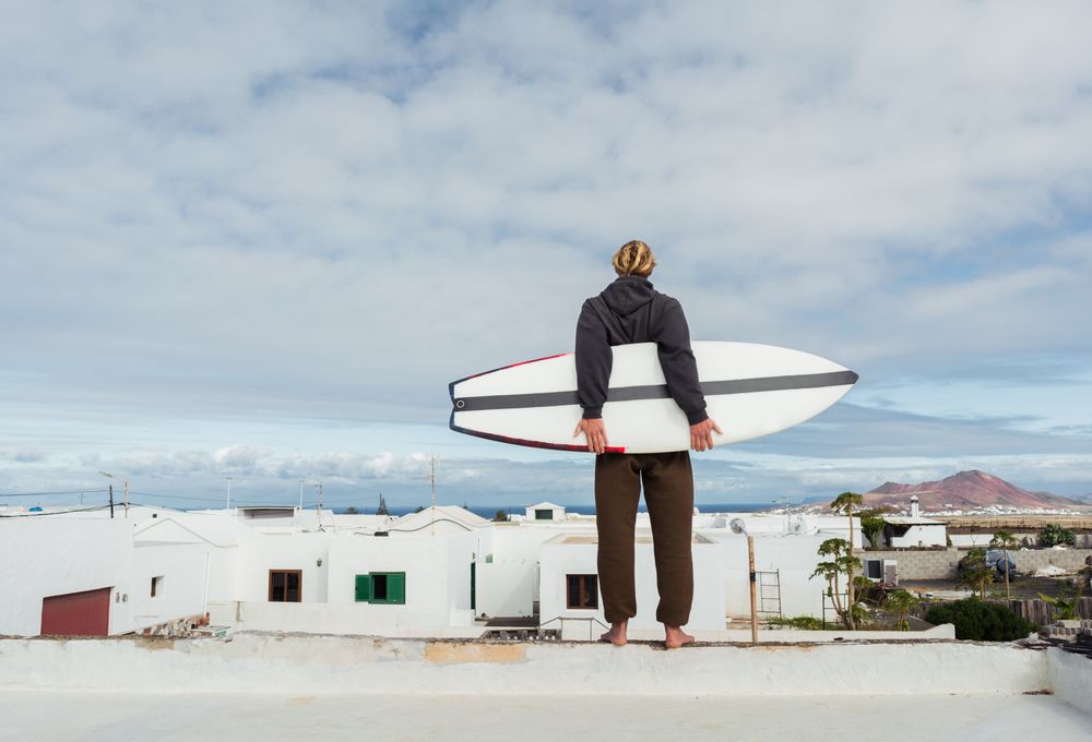 Man standing with surf board on roof of building and looking at mountain